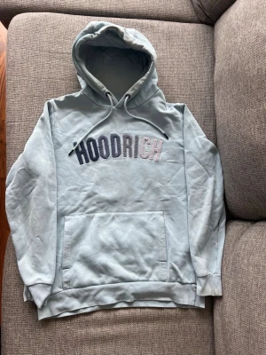 Ljusblå HOODRICH hoodie med huva - Snygg ljusblå hoodie från HOODRICH med stor magficka och broderad logga i svart, grått och vitt på bröstet. Klassisk huva med dragsko och ribbade muddar vid ärmar och nederkant. Perfekt för en chill och avslappnad stil.