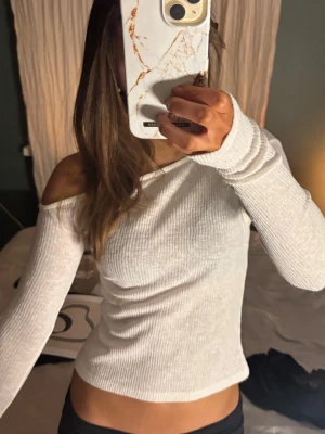 Vit ribbad offshoulder topp - Trendig vit ribbad topp med offshoulder-design och långa ärmar. Sitter tight och har en snygg, enkel look som passar perfekt till jeans eller kjol. Materialet känns mjukt och stretchigt, vilket gör den bekväm att bära hela dagen.