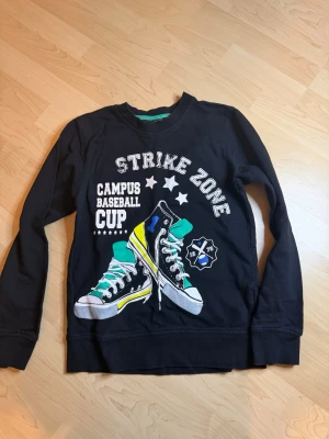 Svart sweatshirt med tryck  - Cool svart sweatshirt från KappAhl med färgglatt tryck av sneakers och texten 'STRIKE ZONE CAMPUS BASEBALL CUP' framtill. Tröjan har rund halsringning med grön detalj och långa ärmar. Perfekt för en avslappnad och sportig stil.