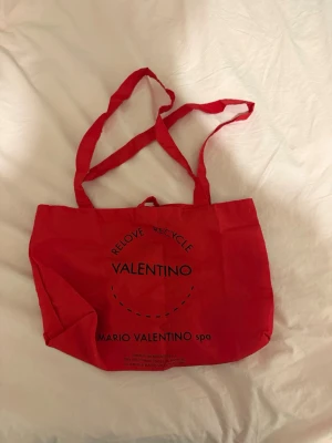 Röd shoppingväska från Valentino - Snygg röd tygkasse från Valentino med tryck i svart text och långa axelband. Perfekt för att bära med sig allt man behöver. Tillverkad i slitstarkt tygmaterial och har en enkel, stilren design med texten 'RELOVE RECYCLE VALENTINO' på framsidan.