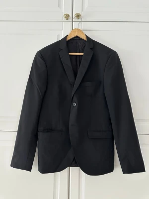 Tiger of Sweden svart ullkavaj slim strl 46-48 fint skick - Snygg svart ullkavaj från Tiger of Sweden – premium slim fit, perfekt för business, fest eller casual med jeans!  •  Material: 96% ull + 4% viskos – håller formen, andas bra, premiumkänsla.  •  Nypris: Ca 3000–4000 kr (kavajdel av kostym ~5000 kr+ totalt).  •  Skick: Mycket fint – inga fläckar/noppor/lukt, använd fåtal gånger. Vänstra armens knappar är lite små trasig amen syns knappast. Mått: Axelbredd 46 cm, ärmlängd 64 cm, midjemått 92 cm – perfekt för kortare/smalare män ca 170–178 cm 