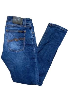 Blå jeans från Nudie Jeans - Säljer ett par klassiska blå jeans från Nudie Jeans med smal passform och snygga slitningar. Jeansen har fem fickor, orange sömmar och en ikonisk vågdetalj på bakfickan. Tillverkade i mjuk denim som sitter skönt hela dagen.