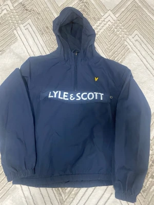 Marinblå hoodie från Lyle & Scott - Snygg marinblå hoodie från Lyle & Scott med stor logga över bröstet och gul broderad fågel på vänster sida. Hoodien har dragkedja framtill, elastiska muddar och är tillverkad i ett mjukt material. Perfekt för en avslappnad och trendig stil. Knappt använd