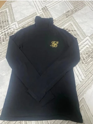 Svart polotröja från SikSilk - Svart långärmad polotröja från SikSilk med gul broderad logga på bröstet. Slim fit modell i mjukt material som sitter snyggt. Perfekt till jeans eller kjol för en clean och stilren look.