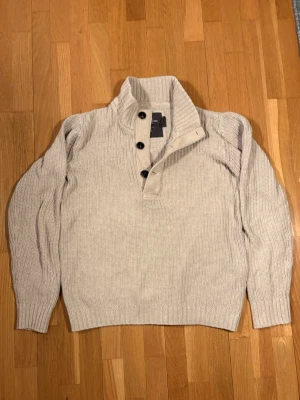 Beige stickad tröja från H&M L.O.G.G. - Säljer en beige stickad tröja från H&M L.O.G.G. med hög krage och fyra svarta knappar framtill. Tröjan har ribbstickade muddar och är gjord i ett grovt stickat material som känns både stilrent och avslappnat.