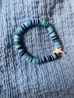 Nelly bracelet💗 - Snyggt armband som är handgjort med platta pärlor i olika blå nyanser och vitt, samt en vit sjöstjärna som detalj. Armbandet har ett randigt mönster och är elastiskt, vilket gör det enkelt att ta på och av. Perfekt för dig som gillar strandvibbar och färgglada accessoarer.