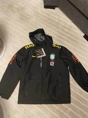 Svart Brasil Nike vindjacka Gore-Tex - Snygg svart vindjacka från Nike med Brasilien-emblem och gula samt orangea loggor. Jackan har huva, dragkedja framtill och två sidofickor. Tillverkad i slitstarkt Gore-Tex-material som står emot regn och vind. Perfekt för dig som gillar sportig stil.