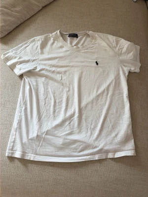 Vit t-shirt från Polo Ralph Lauren - Klassisk vit t-shirt från Polo Ralph Lauren med rund hals och korta ärmar. Diskret mörkblå logga broderad på bröstet. Tillverkad i mjuk bomull för en skön och avslappnad stil.