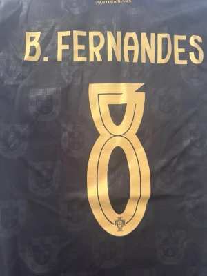 Portugal tröja B. Fernandes Puma M - Snygg svart Portugal fotbollströja från Puma med guldiga detaljer, nummer 8 och B. Fernandes på ryggen. Tröjan har diskreta sköldmönster i tyget och en klassisk krage med rödgrön rand. Tillverkad i återvunnet material (Re:Fibre). Perfekt för match eller träning.