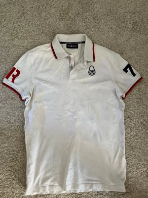 Vit pikétröja från sail racing  - Vit pikétröja från sail racing med röda och svarta detaljer på ärmarna och kragen. Tröjan har korta ärmar, knappslå och broderad logga på bröstet. Perfekt för en sportig och stilren look.