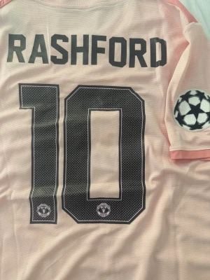 Manchester United Rashford tröja - Ljusrosa fotbollströja med Rashford och nummer 10 på ryggen. Tröjan har svarta detaljer, Manchester United-logga på siffrorna och Champions League-märke på ärmen. Tillverkad i lätt och ventilerande material, perfekt för träning eller match.