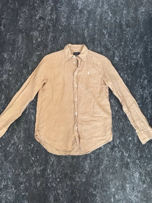 Beige Polo Ralph Lauren Skjorta - Snygg linneskjorta från Polo Ralph Lauren i en varm, cognac-aktig beige ton. Klassisk modell med relaxed fit, avslagen krage och den ikoniska broderade ponnyn på bröstet. Linnetyget ger den där äkta, avslappnade linneskrynklan som bara blir snyggare ju mer den bärs – perfekt att styla både uppknäppt över en tee eller helt knäppt till chinos.