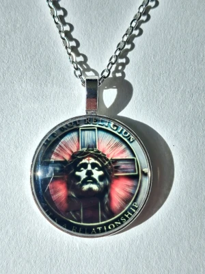 Halsband med Jesusmotiv och kors - Runt halsband med silverfärgad kedja och hänge med motiv av Jesus framför ett kors. Texten runt motivet lyder 'It's not religion, it's a relationship'. Hänget har en glansig yta i 2D bakgrunden är svart med röda och vita detaljer. Materialet rostfritt stål 