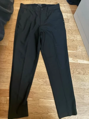 Svarta kostymbyxor från Jack&Jones - Snygga svarta kostymbyxor från Jack&Jones med klassisk rak passform och pressveck. Byxorna har bälteshällor, knäppning fram och två bakfickor med knapp. Perfekta för en stilren look.