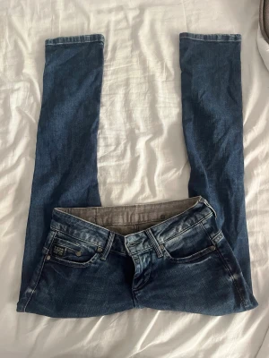 Blå slim fit jeans från G-Star RAW - Säljer ett par blå slim fit jeans från G-Star RAW med klassisk femficksdesign och snygga slitningar. Jeansen har låg midja, dragkedjegylf och detaljerade sömmar. Perfekta för en avslappnad och trendig look. 25/30 innebenslängden 73 midjemåttet 35