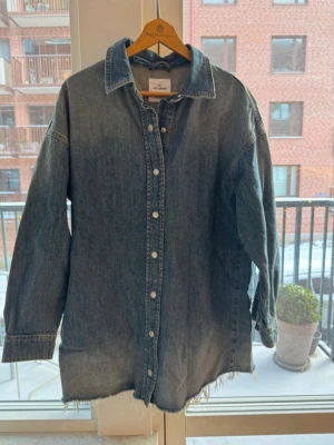 Oversized jeansskjorta/klänning från Pull&Bear - Säljer en oversized jeansskjorta/klänning från Pull&Bear i klassisk blå denim. Skjortan har långa ärmar, knappar framtill och rå, fransad nederkant för en avslappnad look. Storlek S/M