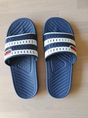 Blåa slides från BLWR med vita ränder 38;or - Säljer ett par blåa slides från BLWR med två vita och blåa ränder över foten. Tillverkade i slitstark plast med räfflad sula för extra grepp. Klassisk design med logga på remmen. St 38. De har aldrig används. 