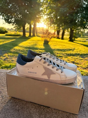 Vita Golden Goose sneakers 🤩 -  Snygga vita sneakers i storlek 43 från Golden Goose med grå stjärndetalj på sidan och mörkblå häl. Skorna har klassisk låg modell, vita snören och svart sula undertill. Perfekta för dig som gillar stilrena och trendiga sneakers med unik design.🤩 