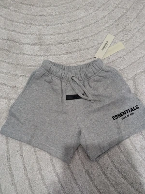Grå Essentials shorts med snörning - Säljer ett par grå shorts från Fear of God Essentials med snörning i midjan och tryckt logga på ena benet. Shortsen har elastisk midja och sidofickor, perfekta för en avslappnad och trendig look. Kids size