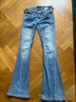 Blå bootcut jeans från True Religion - Säljer ett par ljusblå bootcut jeans från True Religion, knappt använda, med klassiska kontrastsömmar och snygga fickdetaljer bak. Jeansen har låg midja, utsvängda ben och är tillverkade i denim med en lätt tvättad look. Perfekta för en avslappnad och trendig stil. I modell Joey, storlek 24 som motsvarar xs.