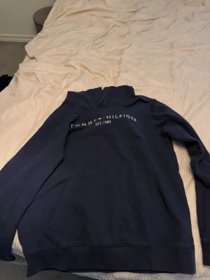 Mörkblå hoodie Tommy Hilfiger - Mörkblå hoodie från Tommy Hilfiger med broderad logga och text 'EST. 1985' på bröstet. Klassisk känguruficka framtill och huva. Andvänd men inga defekter och har används väl skött 