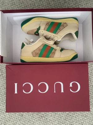 Gucci sneakers med gröna och röda ränder - Snygga sneakers från Gucci i beige och grönt med klassiska gröna och röda ränder på sidan. Skorna har detaljer i mocka och canvas, samt grön yttersula med bi-mönster. Perfekta för dig som vill ha en ikonisk och trendig look.