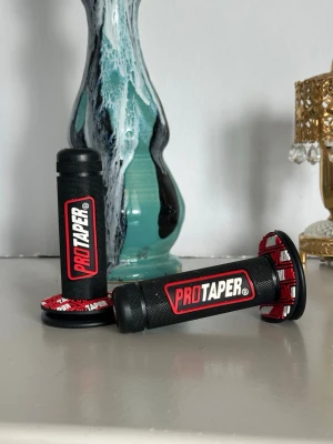 ProTaper svarta handtag till cross - Säljer ett par svarta handtag från ProTaper med röd och vit detalj vid ändarna. Handtagen är tillverkade i gummi och har ett greppvänligt mönster samt tydlig ProTaper-logga i rött och vitt. Perfekt för cross eller motocrossstyre.