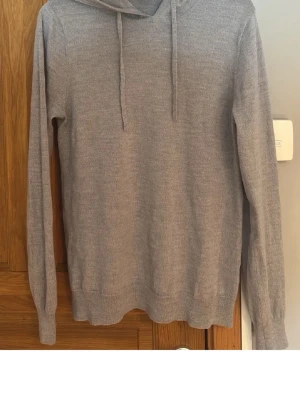 Ljusgrå hoodie i tunn stickad bomull - Säljer en ljusgrå hoodie med huva och dragsnören. Hoodien är tunnstickad och har långa ärmar samt ribbade muddar vid ärmslut och nederkant. Perfekt för dig som gillar en enkel och stilren look.