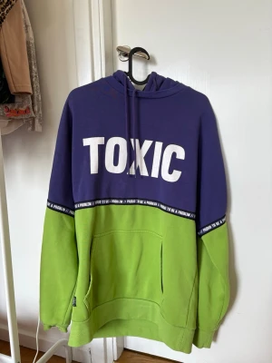 Lila och grön hoodie med TOXIC-tryck - Cool hoodie i lila och grönt med stort vitt TOXIC-tryck på bröstet. Har huva med dragsko och magficka. Svart band med texten 'Proud to be a problem' längs bröstet. 