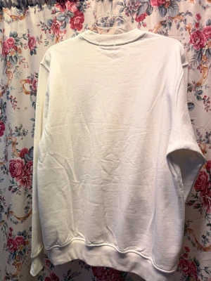 Vit sweatshirt Prada  - Säljer en clean vit sweatshirt från Prada med klassisk rund hals och ribbade muddar. Tröjan är tillverkad i mjuk bomull och har en diskret Prada-logga i metall på bröstet. Perfekt för dig som gillar exklusiva och minimalistiska plagg.