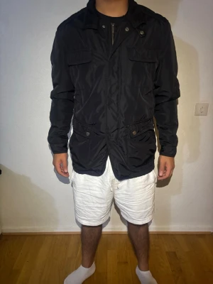 Svart massimo dutti vindjacka  - Svart vindjacka med hög krage och flera praktiska fickor framtill. Jackan har lång ärm, knappar och dragkedja framtill samt en loose passform. Perfekt för dig som vill ha en enkel och stilren jacka till vardags.