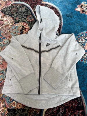 Grå zip hoodie från Nike - Snygg grå hoodie från Nike med dragkedja framtill och svart Nike-logga på bröstet. Hoodien har en huva, svarta detaljer längs dragkedjan och en vågig svart linje över bröstet. Tillverkad i mjukt sweatshirtmaterial och har praktiska fickor på sidorna.