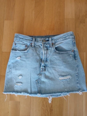  jeanskjol från Levi's strl 27 - Säljer en ljusblå jeanskjol från Levi's med rå kant nertill och slitna detaljer framtill. Kjolen har klassiska fem fickor, bälteshällor och Levi's-lapp bak i midjan. Nypris 750 kr