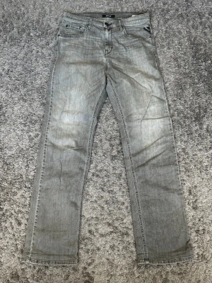 Grå jeans från Replay - Säljer ett par grå jeans från Replay med klassisk femficksmodell och raka ben. Jeansen har en snygg tvättad look och Replay-logga på myntfickan. Tillverkade i mjuk denim som ger en skön känsla och passar till många olika stilar.