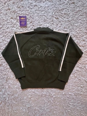 CORTZ- Zip up - Mörkgrön hoodie från Crutz med dragkedja och vita ränder längs ärmarna. Broderad logga på bröstet och stor grafisk text på ryggen. Tillverkad i mjukt material, ribbade muddar och hög krage för extra stilpoäng.