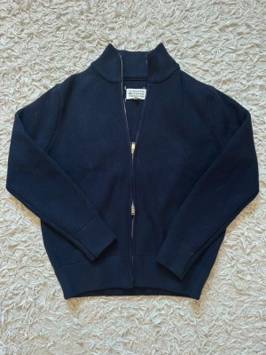  Maison Margiela- heavy Zip - Snygg svart ribbstickad ziptröja från Maison Margiela med hög krage och tvåvägsdragkedja. Tröjan har långa ärmar, diskreta sömmar och de ikoniska fyra vita stygn på ryggen. Perfekt för dig som gillar minimalistisk och stilren design. Tjock och varm! 