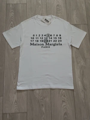 Vit Maison Margiela t-shirt med tryck - Vit t-shirt från Maison Margiela med ikoniskt svart tryck framtill och rund halsringning. Modellen är klassisk och har korta ärmar. Perfekt för dig som gillar stilrena och trendiga plagg med designer-touch.