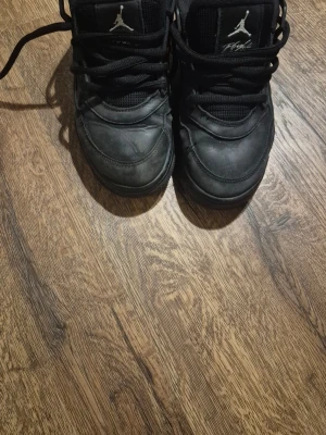 Svarta AIR 4 RM BG SNEAKERS - Säljer ett par helsvarta Air Jordan sneakers med klassisk siluett och Jumpman-logga på plösen. Skorna har en robust sula, svarta skosnören och detaljer i både läder och mesh för en sportig look. Perfekta för dig som gillar streetwear och vill ha något stilrent.