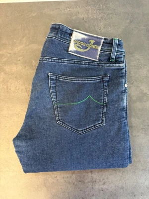 Blå jeans från Jacob Cohën W35 fodrade  - Snygga blå jeans från Jacob Cohën med klassisk femficksmodell och grön detaljsöm på bakfickan. Jeansen har rak passform och är tillverkade i mjukt denimtyg. Perfekta för dig som gillar stilrena och tidlösa jeans med en twist.