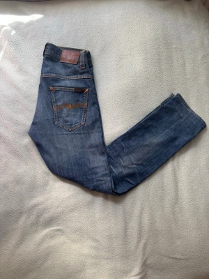 Slimfit Nudie Jeans - Säljer ett par klassiska blå jeans från Nudie Jeans med snygga slitningar/fades och kontrastsömmar. Modellen har raka ben och två bakfickor med den ikoniska Nudie-bågen i orange. Perfekta för en avslappnad och trendig look. Storlek W30 L34, men sitter som W30 L32. Osäker på modell, men tror det är Lean Dean. Tveka inte att skriva om frågor uppstår och priset går att förhandla