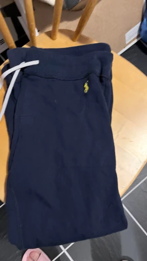 Mörkblå mjukisbyxor från Polo Ralph Lauren - Snygga mörkblå mjukisbyxor från Polo Ralph Lauren med klassisk gul logga på vänster sida. Byxorna har ribbade muddar vid bensluten, dragsko i midjan och mjukt bomullsmaterial. Perfekta för chill dagar eller när du vill vara bekväm.