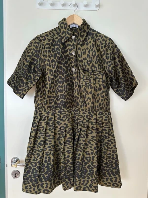 Leopardmönstrad kortklänning från Ganni - Cool kortklänning från Ganni i leopardmönster med glittriga knappar framtill. Klänningen har skjortkrage, korta ärmar och en bröstficka. Materialet känns som en blandning av polyester och bomull, perfekt för att sticka ut.