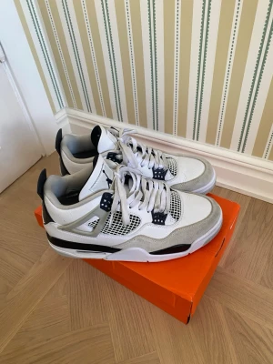 Air Jordan 4 Retro vit/svart/grå - Snygga Air Jordan 4 Retro sneakers i vitt, svart och grått med meshpaneler och mockadetaljer. Klassisk Jumpman-logga på hälen och tungans framsida. Skorna har en chunky sula och coola plastdetaljer vid snörningen.