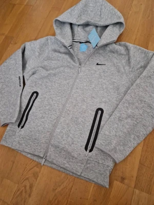 Grå Nike NOCTA hoodie med dragkedja - Snygg grå hoodie från Nike NOCTA med dragkedja framtill och två svarta fickor med dragkedja. Klassisk huva och diskret Nike-logga på bröstet. Tillverkad i mjukt material som passar perfekt till en avslappnad streetwear-look.