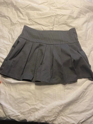 Grå rutig kjol från H&M - Grå kortkjol från H&M med rutigt mönster och bred linning. Kjolen har veckad design som ger en snygg volym och klassisk look. Perfekt att styla med en enkel topp eller stickad tröja.