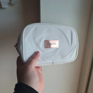 Lancôme necessär vit - Snygg vit necessär från Lancôme i tygmaterial med en rektangulär form. Framsidan har en metallbricka med Lancôme-loggan i guld. Perfekt för att förvara smink eller småsaker. Enkel och stilren design som är lätt att ta med sig. Kan finnas spår av användning.