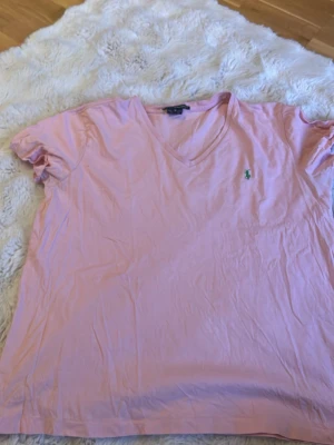 Rosa v-ringad t-shirt från Polo Ralph Lauren - Snygg rosa t-shirt från Polo Ralph Lauren med klassisk grön broderad logga på bröstet. T-shirten har v-ringning och korta ärmar, perfekt för en avslappnad och stilren look. Tillverkad i mjuk bomull för maximal komfort.