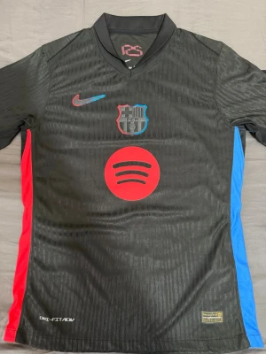 Svart FC Barcelona matchtröja Nike - Säljer en svart FC Barcelona matchtröja från Nike med Dri-FIT ADV-material. Tröjan har klubbmärke och Nike-logga på bröstet, Spotify-logga i rött på magen samt röda och blå detaljer på sidorna. Perfekt för dig som älskar fotboll och vill ha en snygg supportertröja.