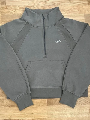 Khaki grön half-zip hoodie från Alo - Snygg khaki grön hoodie från Alo med half-zip dragkedja och hög krage. Tröjan har en stor magficka och ribbade muddar vid ärmslut och midja. Tillverkad i mjukt material, perfekt för chill eller träning. Minimalistisk design med Alo-logga på bröstet.