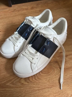 Vita sneakers med marinblå  - Snygga vita sneakers från Valentino med bred marinblå läderdetalj över snörningen och vita skosnören. Skorna har en låg profil och coola nitar på hälen samt en robust sula. Helt nya använda endast EN GÅNG. Storlek 39
